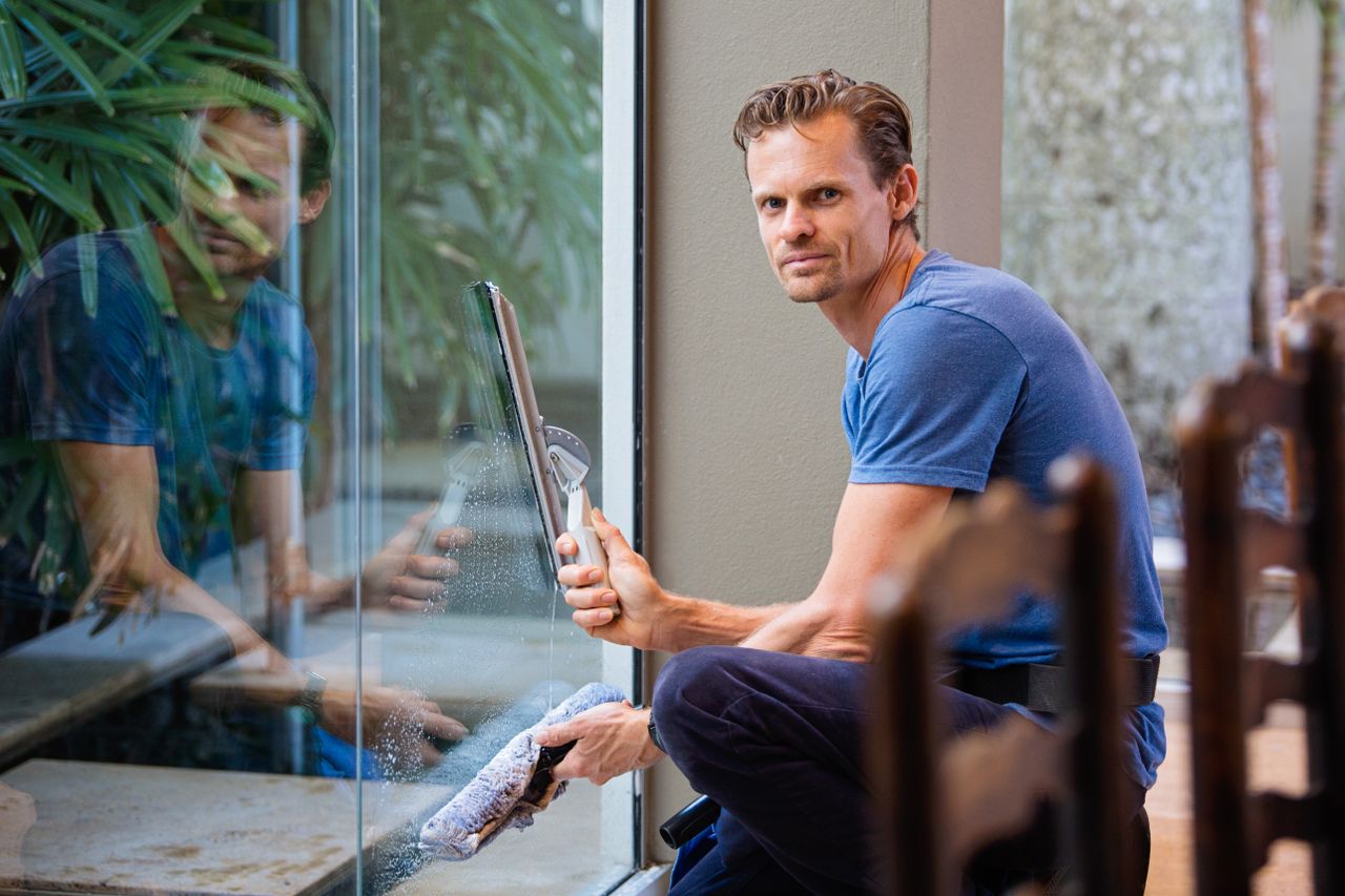 services-06 A man in a blue t-shirt cleaning large glass pane with a squeegee indoors.